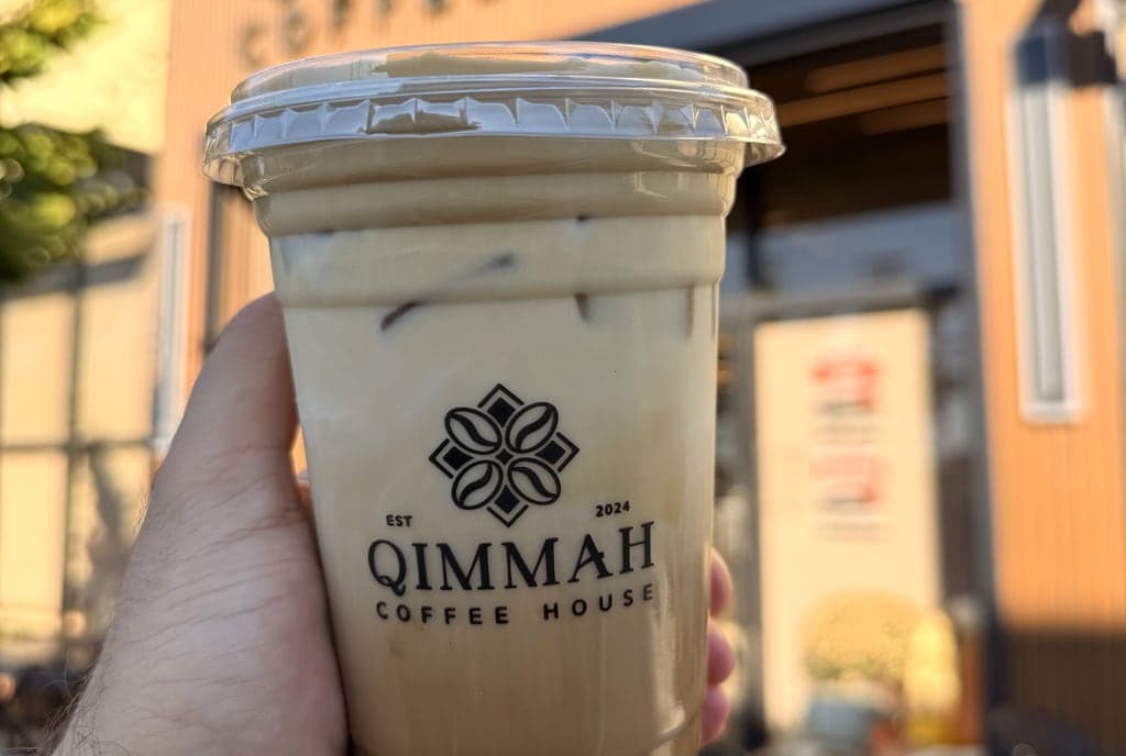 An iced salted maple brown butter latte in a Qimmah Coffee House cup, held up outside the shop