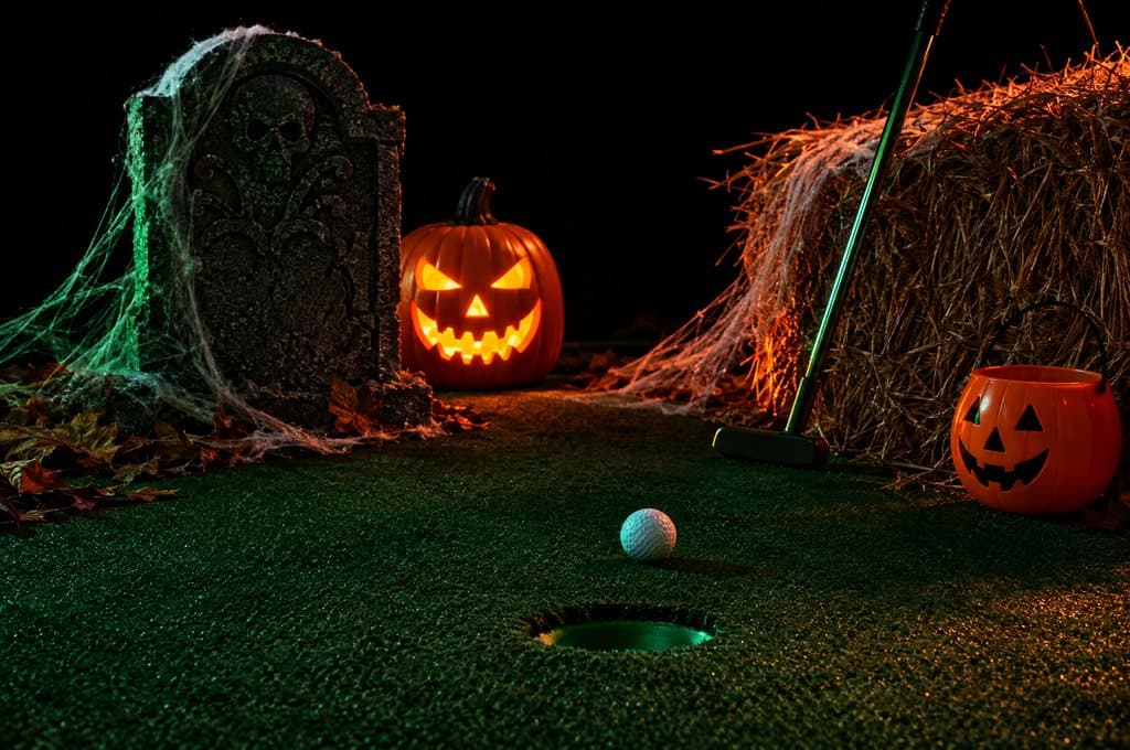 A spooky mini golf hole with a tombstone, jack-o'-lanterns and a putter